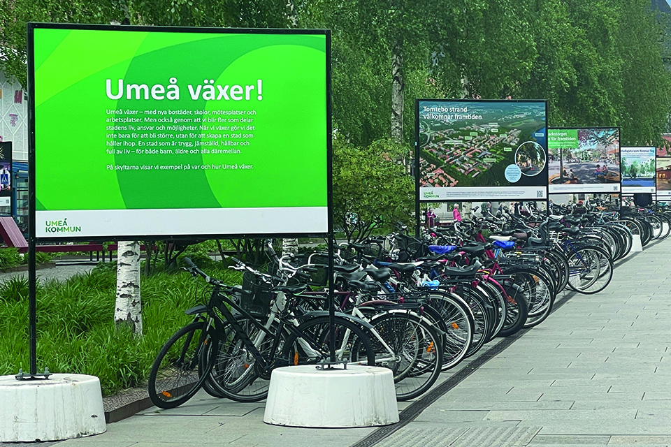 Informationsskyltar från Umeå kommun står uppställda längs en gångväg intill en rad cykelställ. Den främsta skylten har rubriken "Umeå växer!" och berättar om stadens utveckling med bostäder, skolor och mötesplatser.
