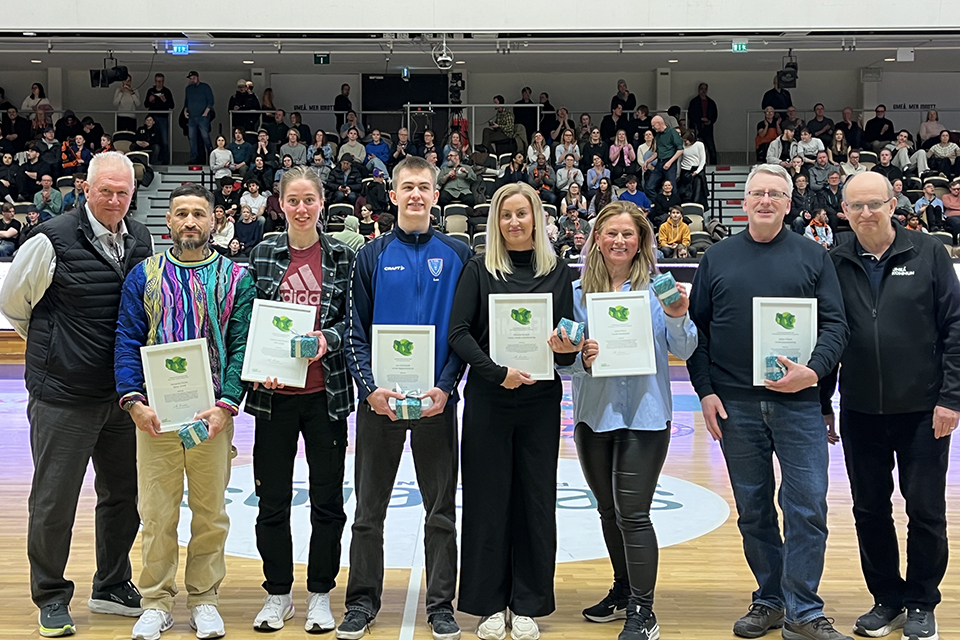 Människor som står på rad mitt i en idrottshall. Sex stycken av personerna håller en tavelram med diplom i händerna.