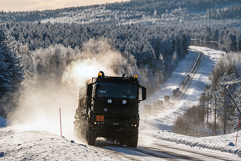 Kolonn med militära lastbilar på väg genom ett vintrigt skogslandskap.