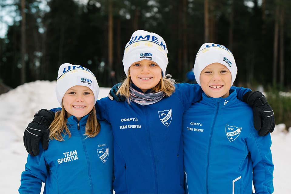 Tre barn poserar framför kamera med en vit SM-mössa på huvudet.