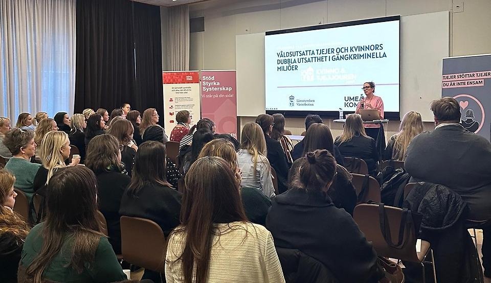 Sal med många åhörare. En kvinna håller ett föredrag framför en Powerpoint-presentation.