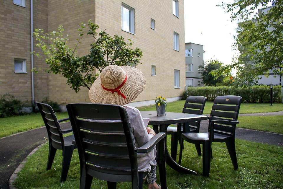 Person med sommarhatt sitter i trädgårdsmöbel på innergård vid flerfamiljshus om sommaren.