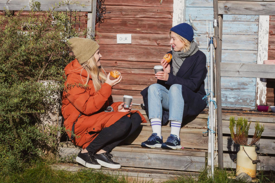 Två unga kvinnor sitter på en trappa utomhus och fikar bulle och kaffe